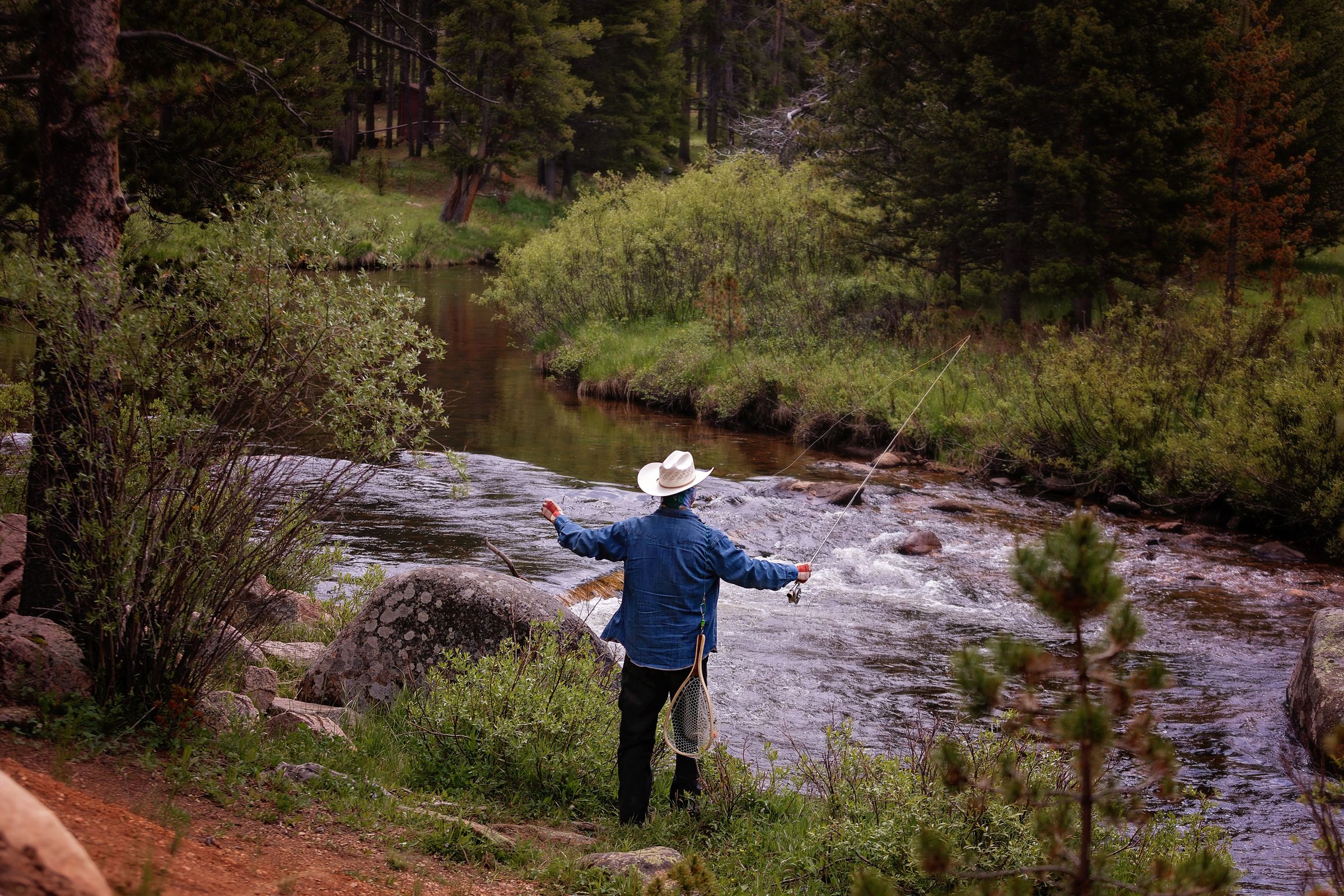 A fly fisherman in a cowboy hat casting into a fast mountain creek